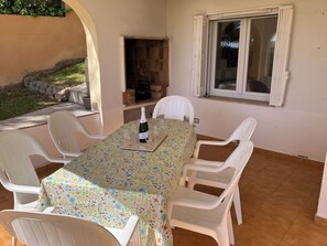 An outdoor patio with a table set for four, surrounded by white plastic chairs, a bottle of champagne, and wine glasses.