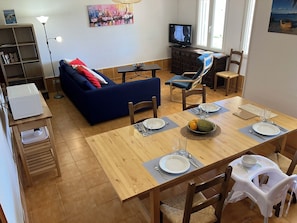 A dining area with a wooden table set for four, a blue sofa, a television, and a bookshelf.