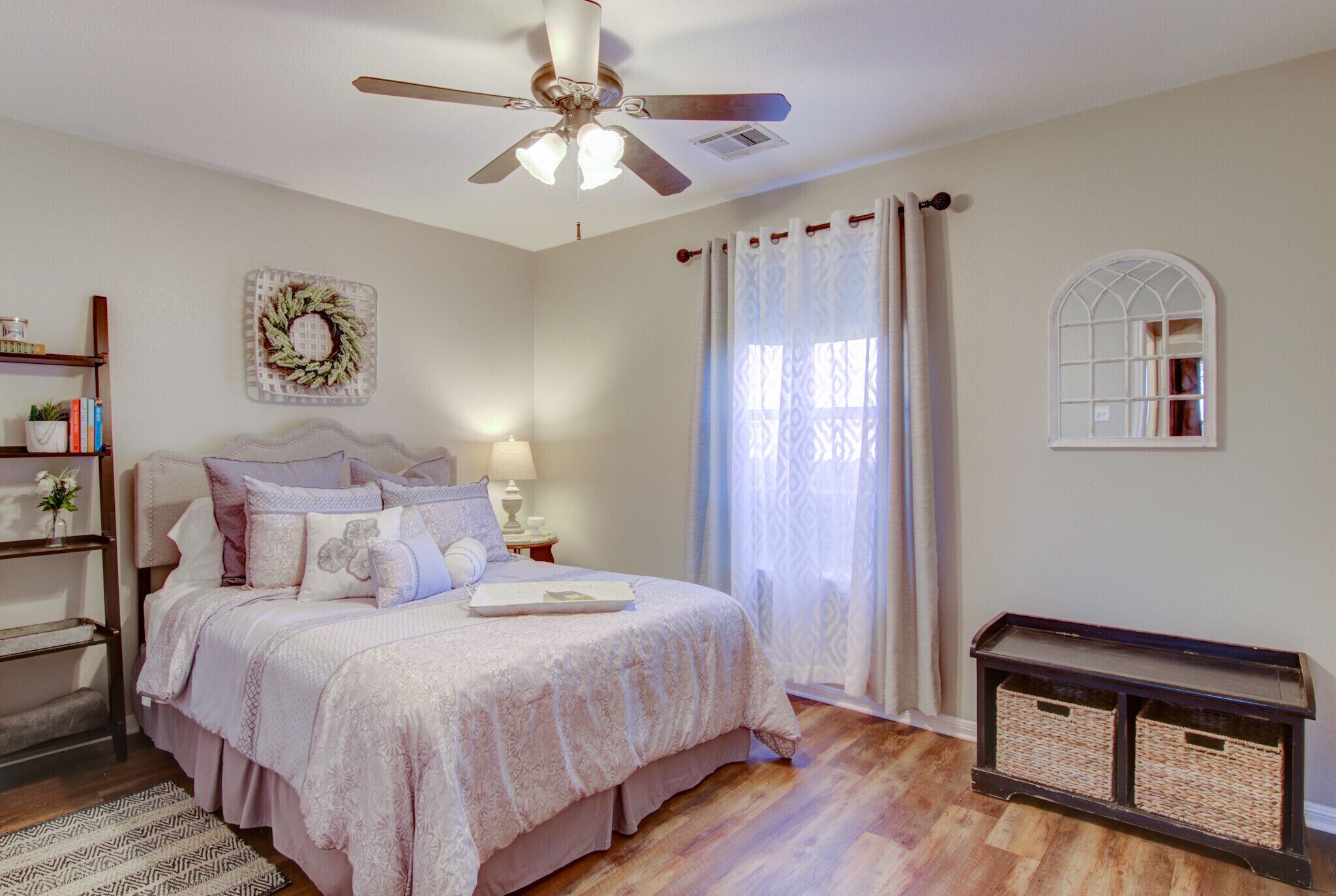 A bedroom with a bed, a ceiling fan, a bedside table, a nightstand, a bookshelf, a decorative wreath, a window with curtains, and a small mirror.