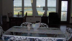 Uma sala de jantar com mesa posta para quatro pessoas, rodeada por cadeiras. Há janelas com persianas, um candelabro e uma porta com painel de vidro.