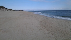 Een breed zandstrand met zachte golven, een heldere lucht en een pier in de verte.