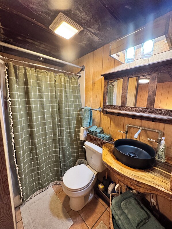 Un baño con detalles de madera, una cortina de ducha a cuadros verdes, un inodoro blanco, un lavabo negro con dispensador de jabón y un espejo sobre el lavabo.