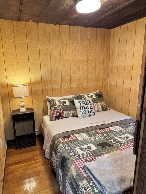 A cozy bedroom with a wooden wall, a bed with patterned bedding, a nightstand with a lamp, and a quilt with a 'Take me to the lake' pillow.