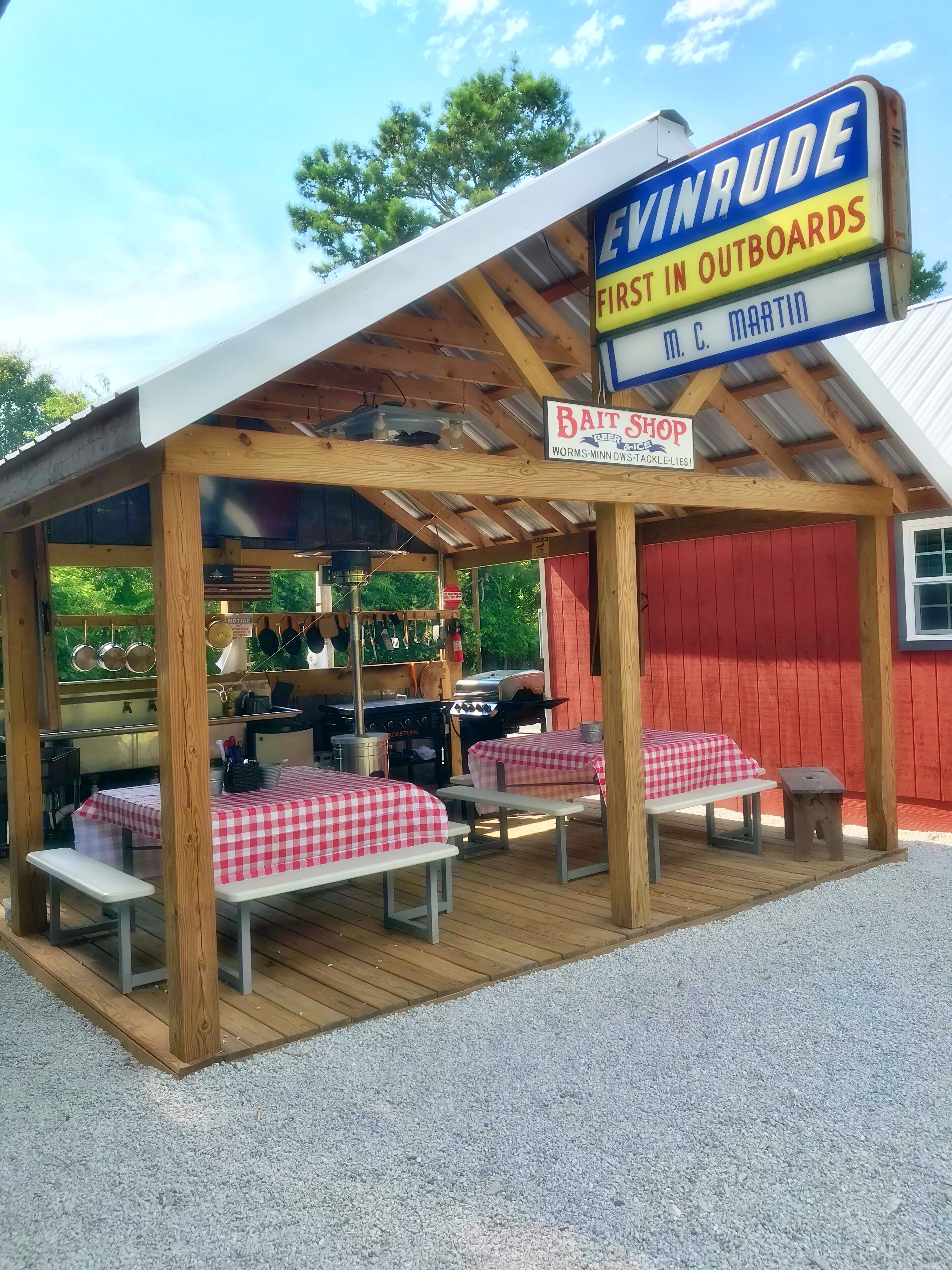 Una estructura de madera al aire libre con un espacio de estar cubierto, una parrilla y un letrero que anuncia 'Bait Shop' y 'Worms-Minn'.