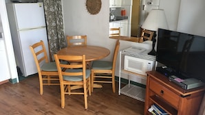 Een compact leefruimte met een houten eettafel, twee stoelen, een magnetron, een televisie en een kitchenette met een koelkast en kastjes.