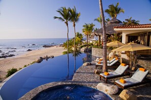 Una piscina infinita con sillones de descanso y sombrillas, una playa y palmeras.