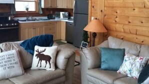 A cozy cabin interior with two beige sofas, a wooden staircase, a kitchen with wooden cabinets, and a small table with a lamp. - Glacier Lake Log Cabin (Oxford)