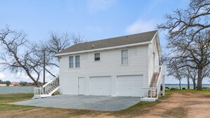 A two-story white house with a front porch, a staircase leading to the garage, and a view of the water in the background. - Small but cozy apartment style lakefront home on Buchanan Dam with great views! (Buchanan Dam)