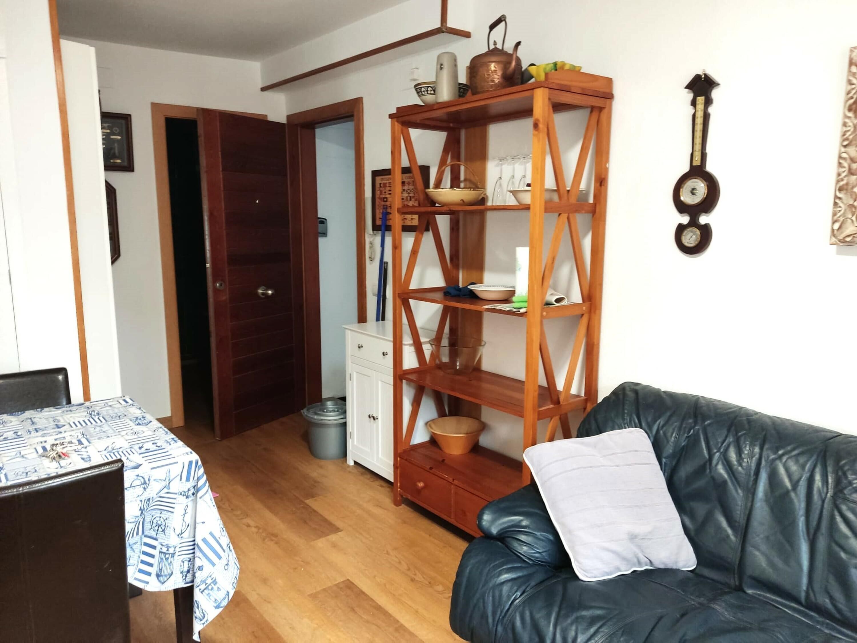 Un salon avec un canapé en cuir noir, une étagère en bois contenant des ustensiles de cuisine, un meuble blanc et une porte en bois donnant sur une autre pièce.