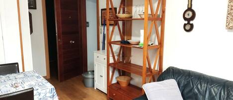 Un salon avec un canapé en cuir noir, une étagère en bois contenant des ustensiles de cuisine, un meuble blanc et une porte en bois donnant sur une autre pièce.