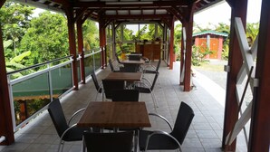 Uma área de estar ao ar livre coberta, com mesas e cadeiras, cercada por uma pérgula de madeira e guarda-corpos de vidro, com vista para um jardim exuberante.