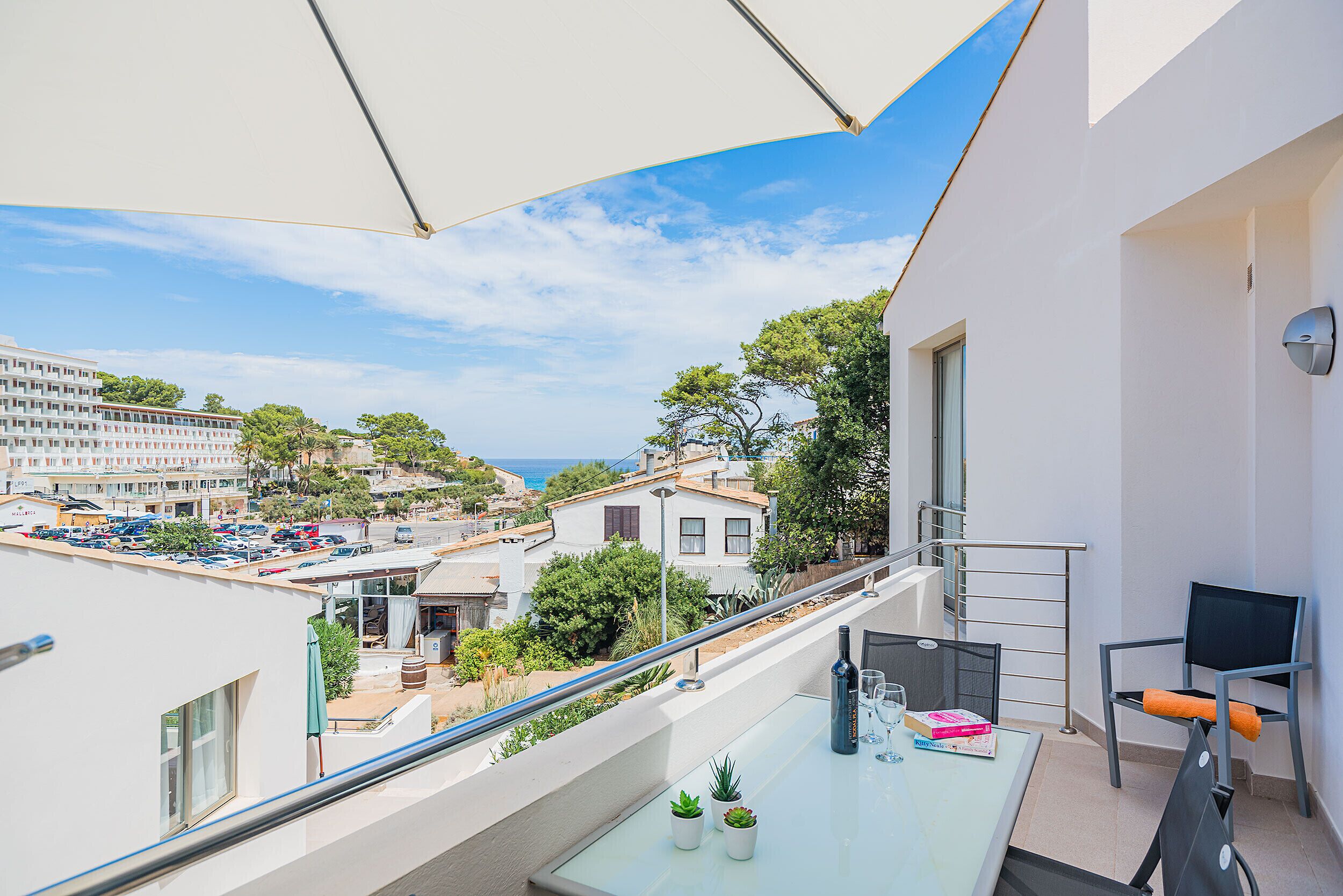 A balcony with a glass table, two potted plants, a chair, and a view of the cityscape and sea.