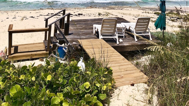 Una playa con una terraza de madera, dos sillas reclinables blancas y un paraguas verde.