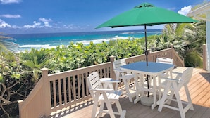 Terraza de madera con una mesa y sillas, con vista a una playa, olas y cielo despejado.