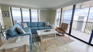 Un salón amplio con un sofá seccional azul, una mesa de centro de madera y un sillón beige. Ventanales grandes ofrecen vista al océano y a un edificio de la calle.