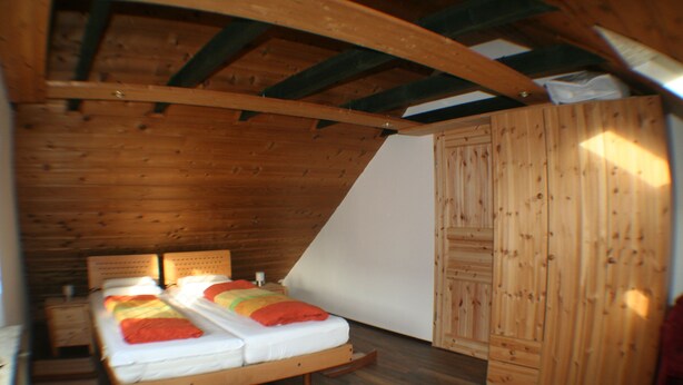 A small, wooden-paneled bedroom with a sloped ceiling, a bed with multiple pillows, and a wooden wardrobe. - Feel-good nest with children's bunk (Dornum OT Dornumersiel)