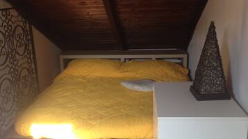 Une chambre avec un plafond en pente, un lit recouvert d’une couverture jaune, une table de chevet blanche et un objet décoratif posé sur la table.