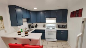 Cocina compacta con armarios azul oscuro, una estufa blanca y un microondas. Hay dos sillas rojas en una pequeña isla.