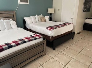 Dos camas con ropa blanca y una manta a cuadros, una mesita de noche con lámpara, y una puerta en una habitación con suelo de baldosas.