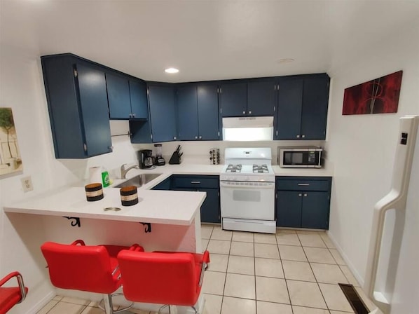 A compact kitchen with dark blue cabinets, a white stove, and a microwave. There are two red chairs at a small island. - 2330/B Comfy 2 bed dual master with Backyard! (San Diego)