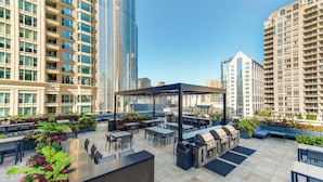 Ein Terrassenbereich im Freien mit Tischen, Stühlen, einem Grill und einer überdachten Sitzgelegenheit, umgeben von hohen Gebäuden.