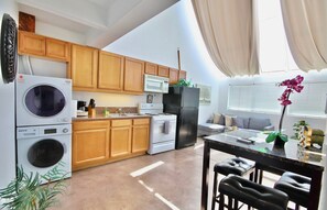 A compact kitchen with wooden cabinets, a washing machine, a microwave, a refrigerator, and a dining area with a table and chairs. - 301 Amazing Sky Loft in Little Italy! (San Diego)