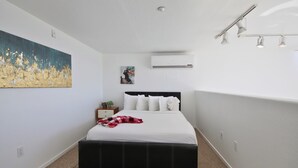 Un dormitorio con cama, mesita de noche, un cuadro, aire acondicionado e iluminación en el techo.