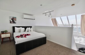 A hotel room with a bed, a nightstand, an air conditioning unit, a window with curtains, and a city view. - 301 Amazing Sky Loft in Little Italy! (San Diego)