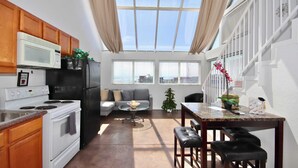 Cocina compacta con microondas, estufa y refrigerador. Cuenta con un área de comedor con mesa y sillas, y una pequeña planta en maceta.
