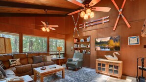 A cozy living room with a leather sofa, a wooden coffee table, and a fireplace. There are ceiling fans, a bookshelf, and a painting on the wall. - (18B) Hiltbrand Hangout at The Redwoods In Yosemite (Yosemite National Park)