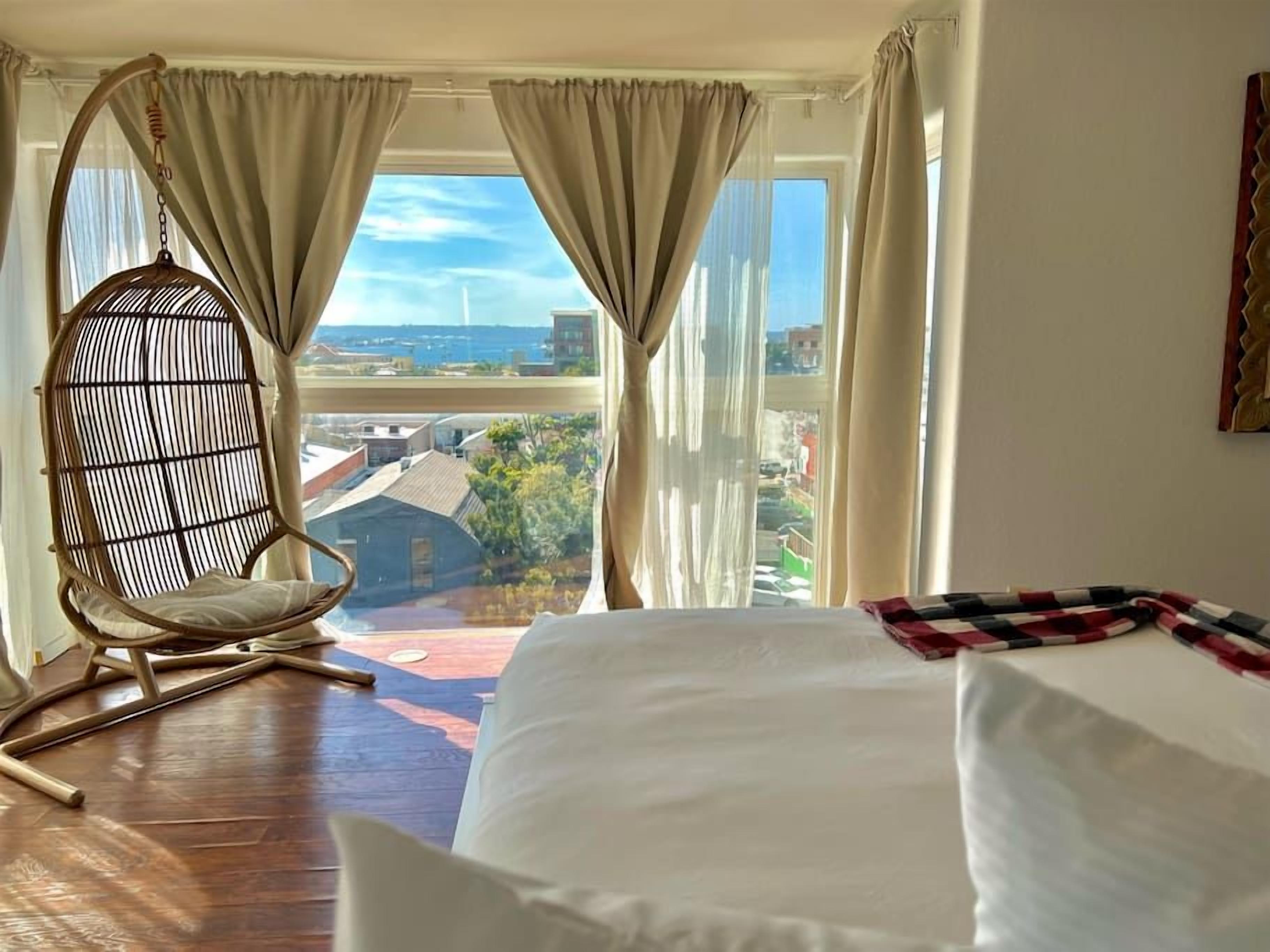 A hotel room with a large bed, a hanging chair, and a view of a cityscape through sheer curtains.