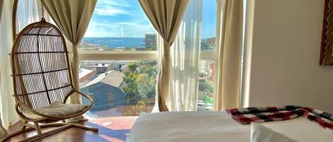 A hotel room with a large bed, a hanging chair, and a view of a cityscape through sheer curtains.