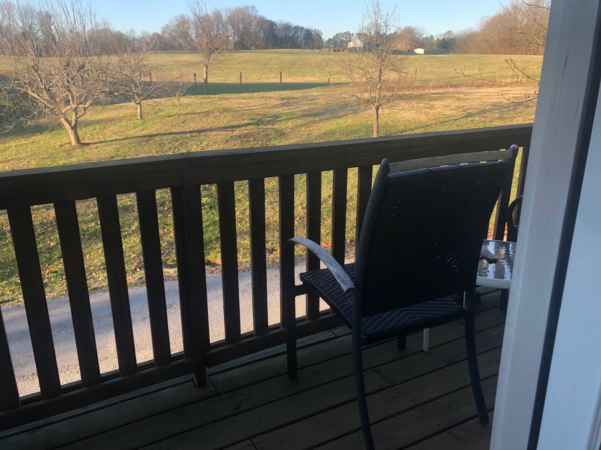 A wooden deck with a chair and table, overlooking a grassy field with trees and a distant house.