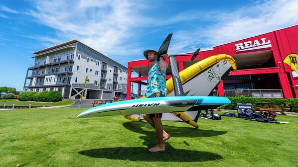 A person holding a surfboard in front of a building with a red and white sign that reads "REAL". - 3-bedroom, 2-bathroom apartment. Walk to beach and access to REAL Watersports (Waves)