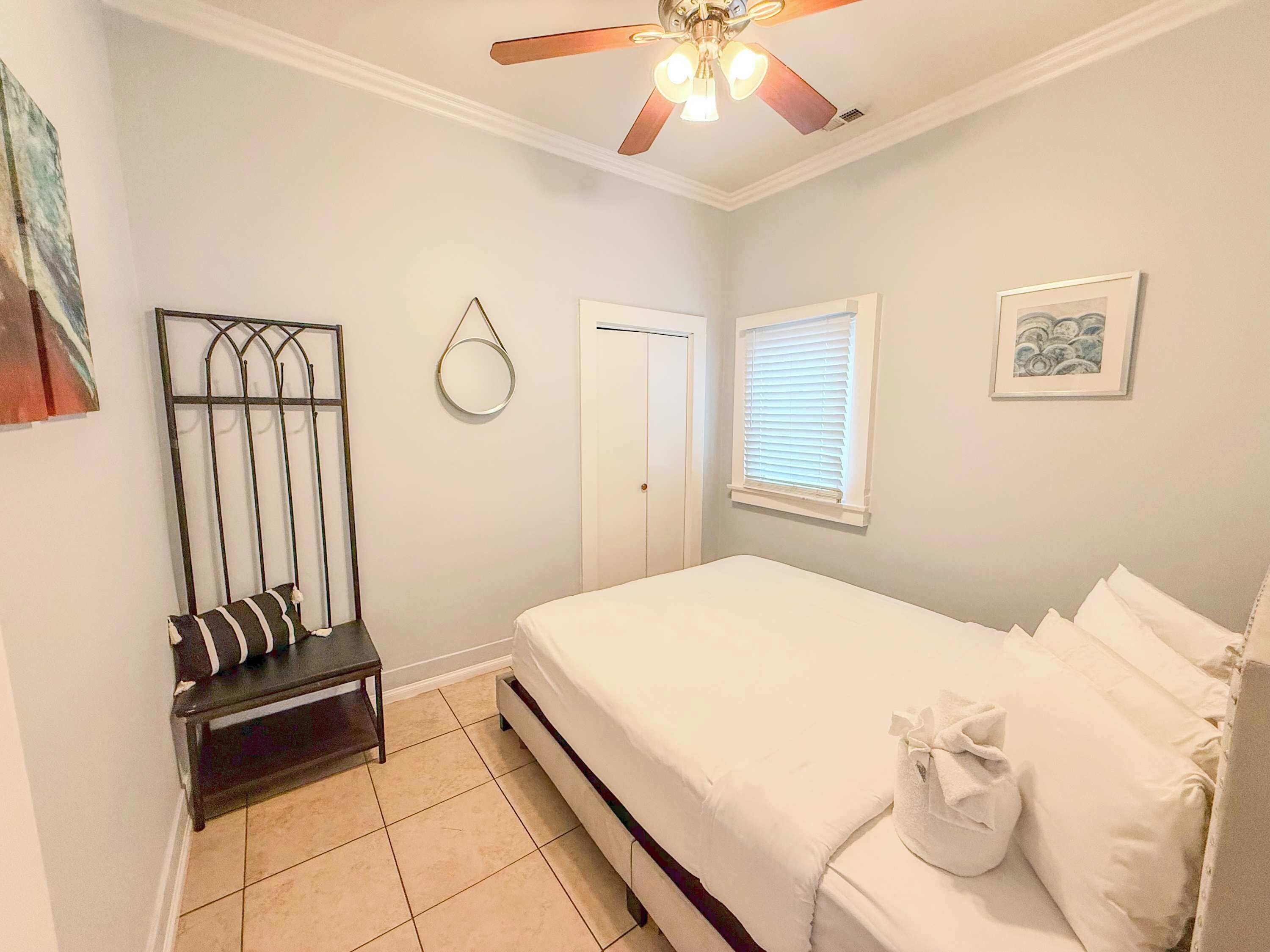 Une chambre à coucher comprenant un lit, un ventilateur de plafond, une fenêtre avec des stores, une porte et une petite table avec une chaise.