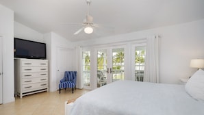 Un dormitorio con una cama grande, un televisor montado en un mueble, una silla y un balcón con vistas a palmeras.