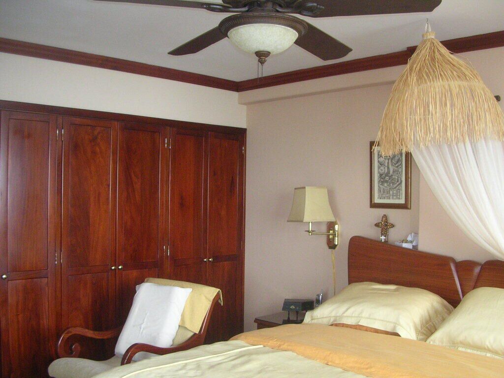 Une chambre d’hôtel comprenant un lit, une chaise, une armoire, une table de chevet, une lampe et un ventilateur de plafond.