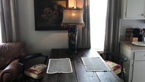 A dining area with a wooden table, two chairs, a table runner, a lamp, a painting, and a kitchen counter with utensils.