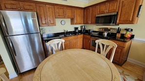 A kitchen with wooden cabinets, a stainless steel refrigerator, a microwave, and a dining table with two chairs. - Pet-Friendly Retreat w/ Private Balcony, Just A Short Walk To The Beach (Tybee Island)