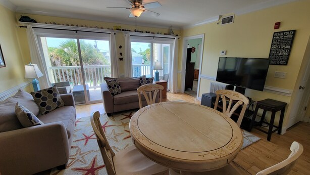 A living room with a round wooden table, two sofas, a TV, and a balcony with a view of palm trees. - Pet-Friendly Retreat w/ Private Balcony, Just A Short Walk To The Beach (Tybee Island)
