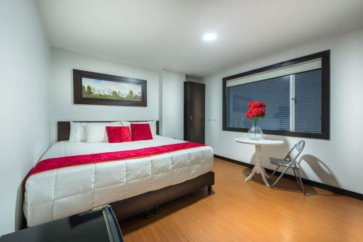 Ein Hotelzimmer mit einem Bett, einem kleinen Tisch mit Blumenvase, einem Stuhl und einem Fenster mit Jalousien.