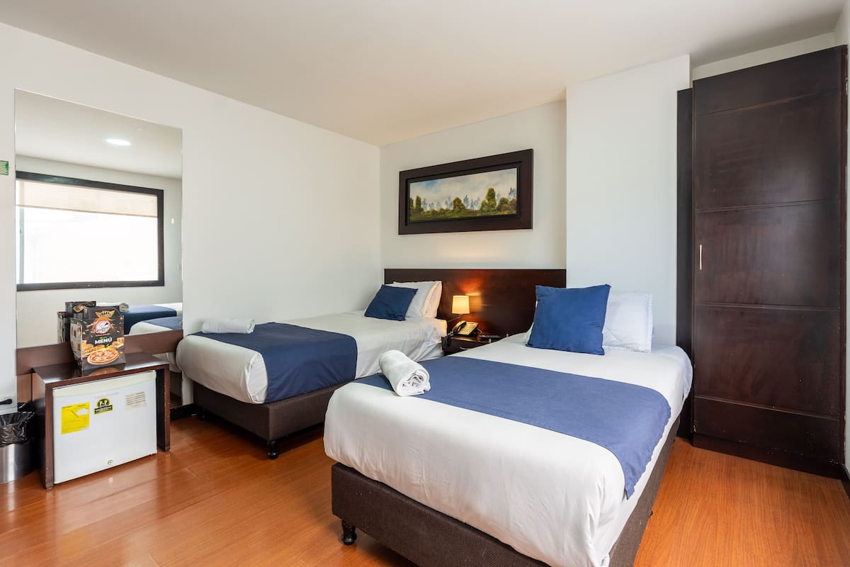 Ein Hotelzimmer mit zwei Betten, einem Schreibtisch mit Speisekartenhalter, einer kleinen Kühlschrank und einem Gemälde an der Wand.