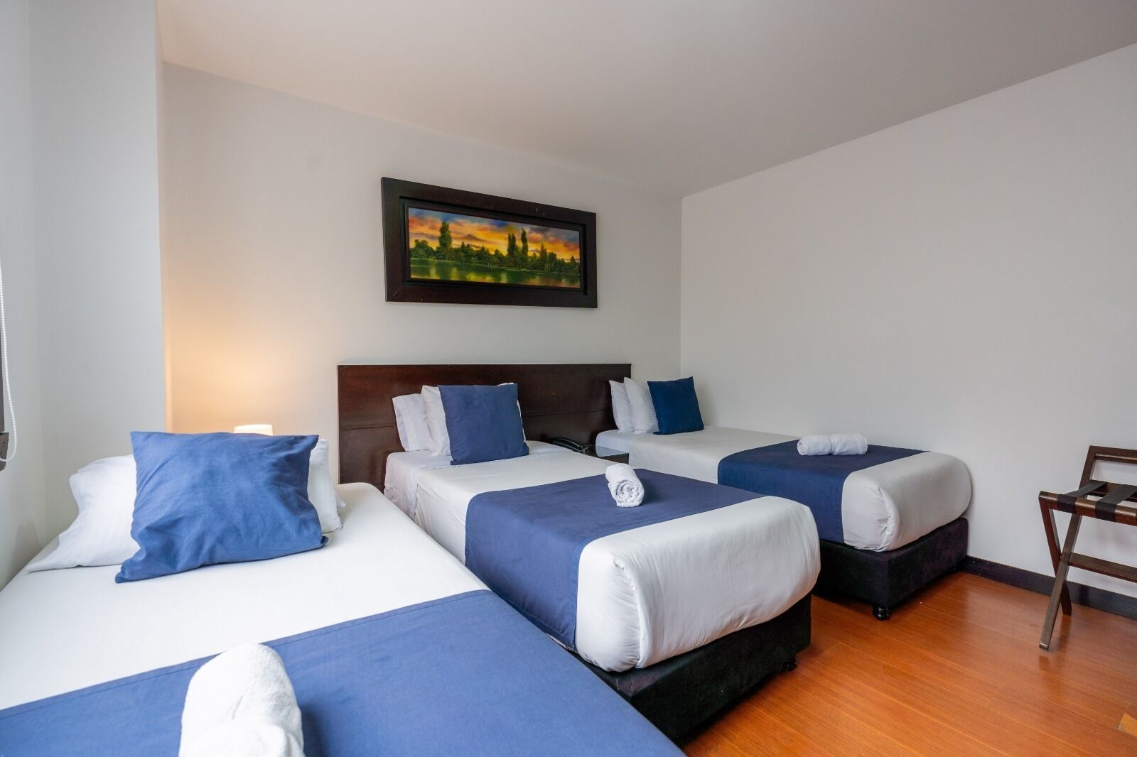 Una habitación de hotel con tres camas, cada una con ropa de cama azul y blanca, cabeceros de madera y un cuadro de un paisaje en la pared.