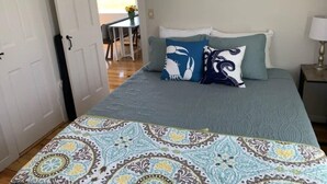 Une chambre à coucher avec un lit, une couvre-lit à motifs et des oreillers. On y trouve une porte qui mène à une autre pièce et une lampe sur une table de chevet.