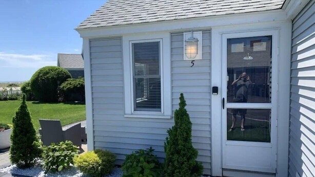 Une petite maison de plain-pied, à l’extérieur blanc, avec une porte d’entrée dotée d’un panneau de verre et le chiffre « 5 » au-dessus de la porte. Devant la maison, on trouve trois petits arbustes coniques, et une allée pavée qui mène à la porte.