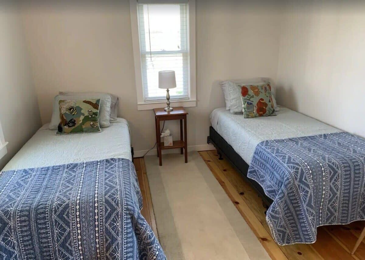 A bedroom with two single beds, each with patterned bedding, a bedside table with a lamp, and a window with blinds.
