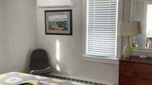 Une chambre à coucher comprenant un lit, une chaise, une commode, une fenêtre avec des stores et un tableau encadré accroché au mur.