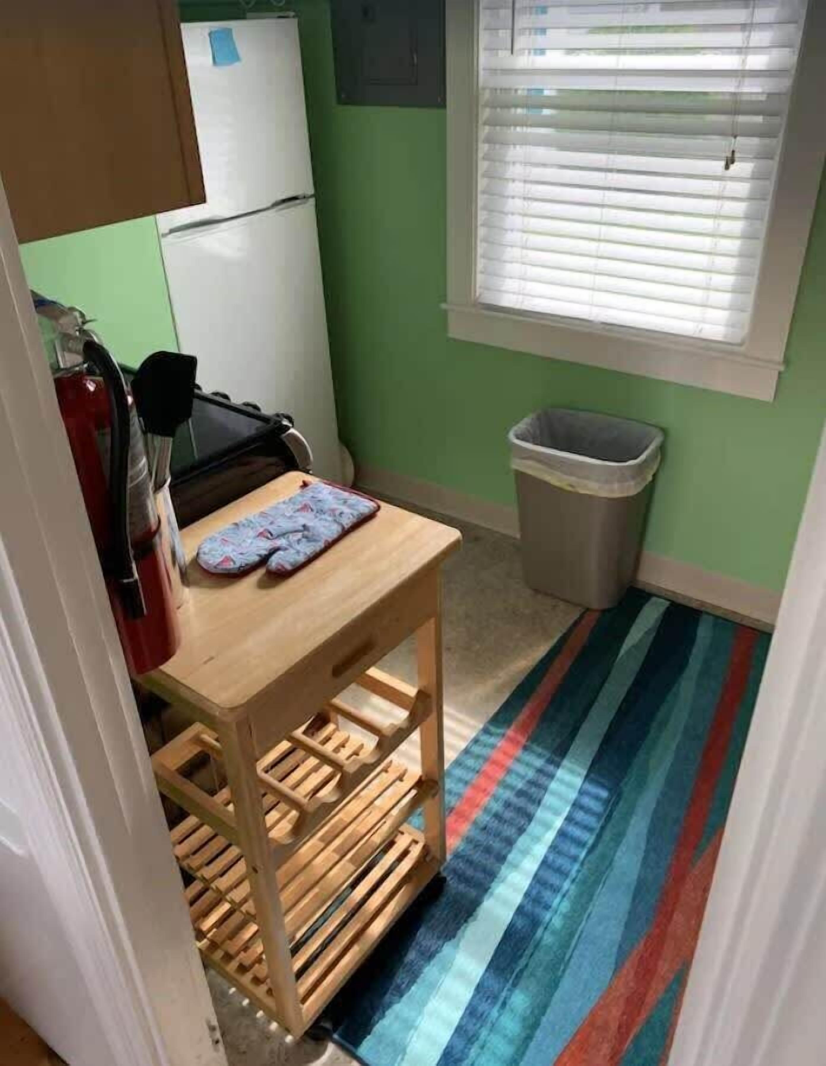 Una pequeña cocina con una mesa de madera, un extintor, un cubo de basura y una ventana con persianas.
