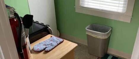Una pequeña cocina con una mesa de madera, un extintor, un cubo de basura y una ventana con persianas.