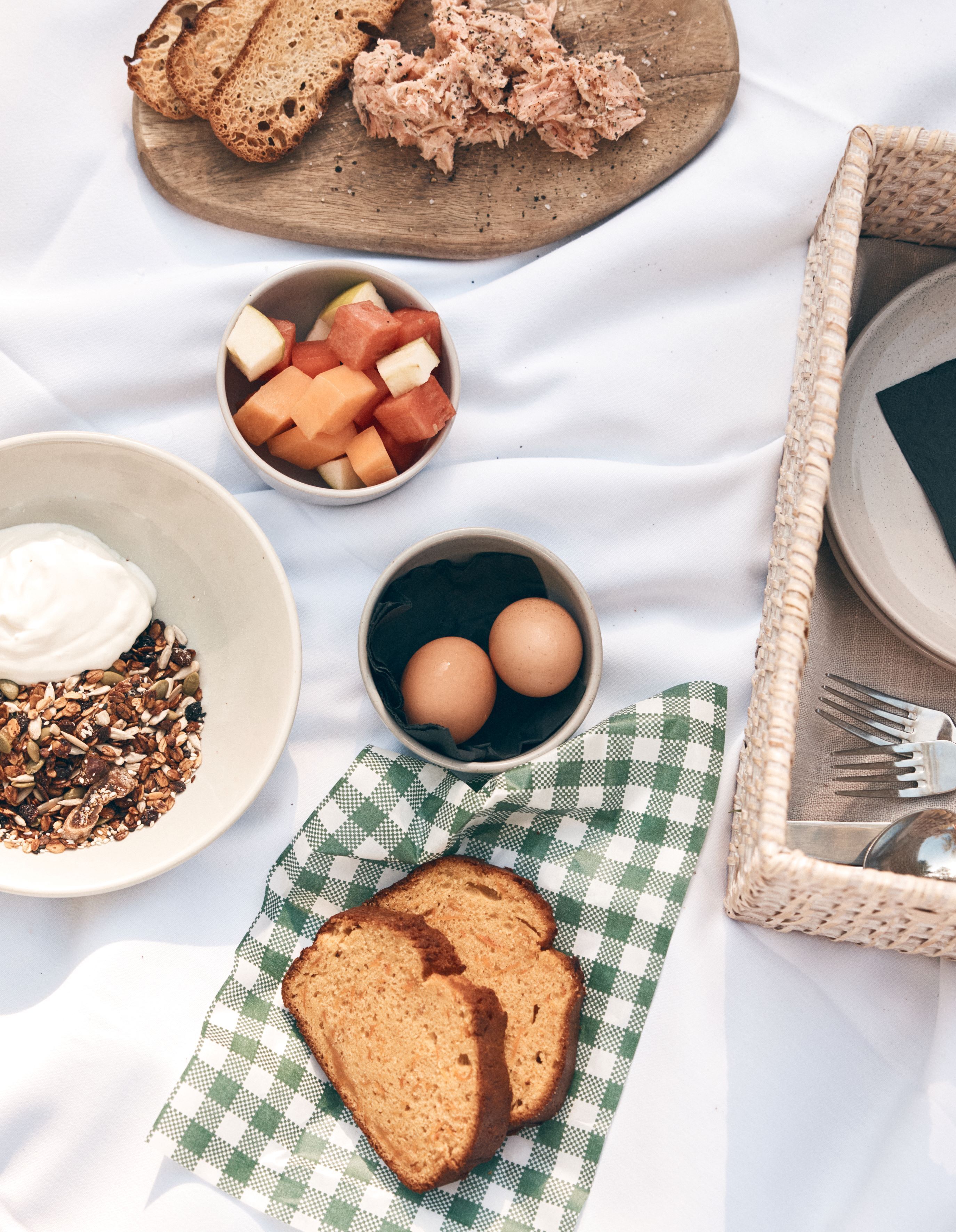 Ontbijt met toast, eieren, yoghurt, granola en meloen op een geruit kleed.